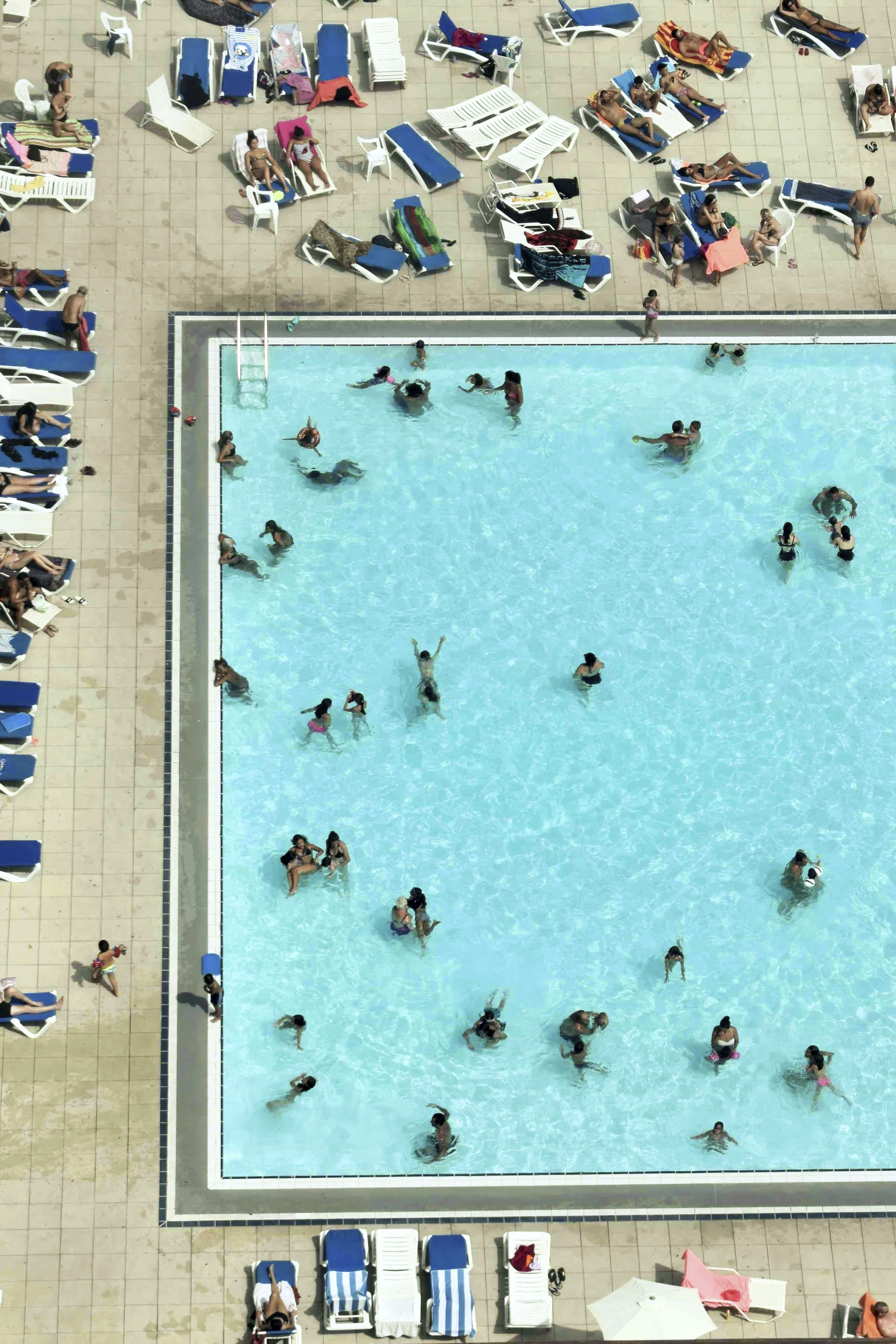 A large outdoor swimming pool seen from above with bathers in the water and people lying on lounge chairs around the pool