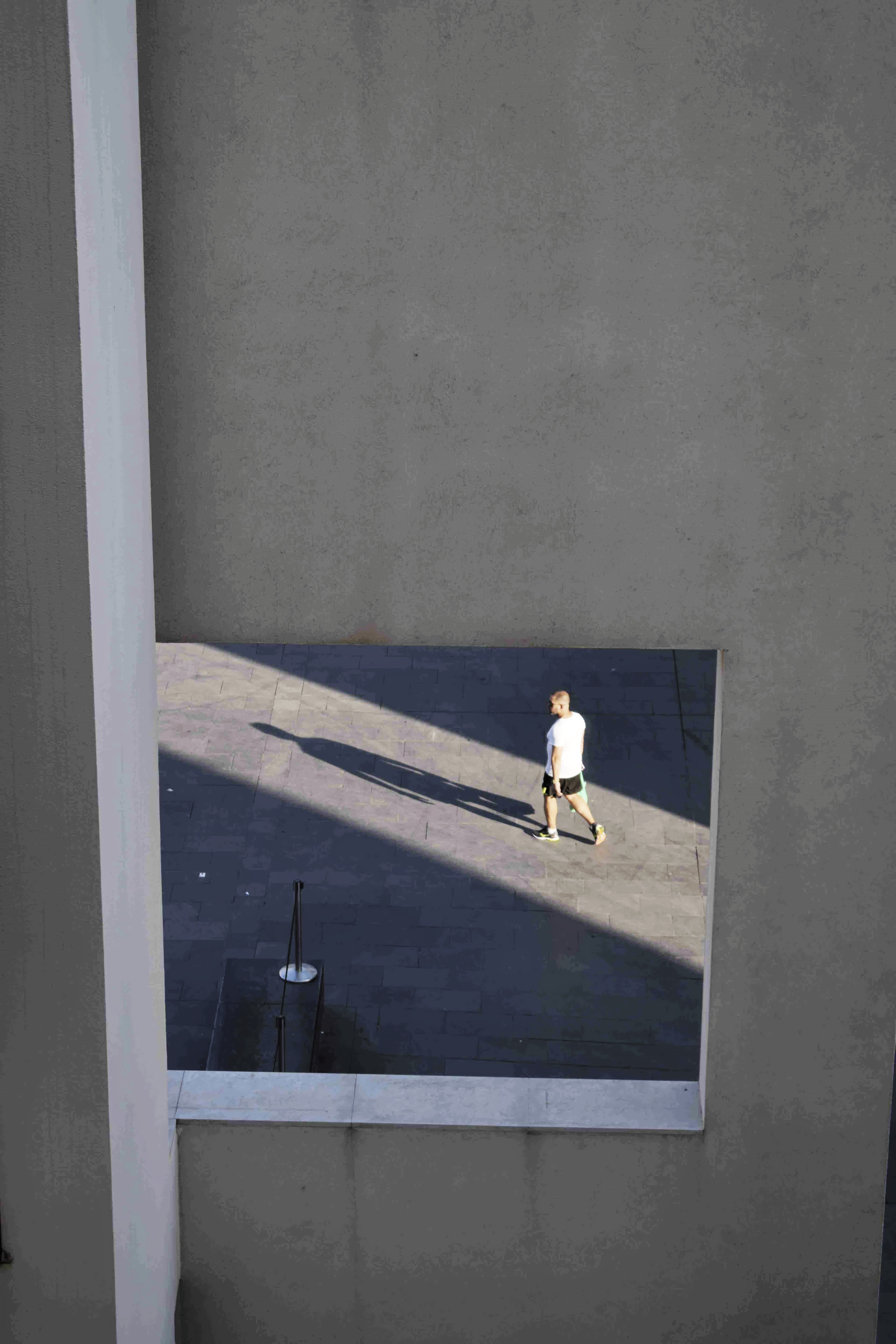  A man walking with his shadow in front of him seen through a square opening in a concrete structure