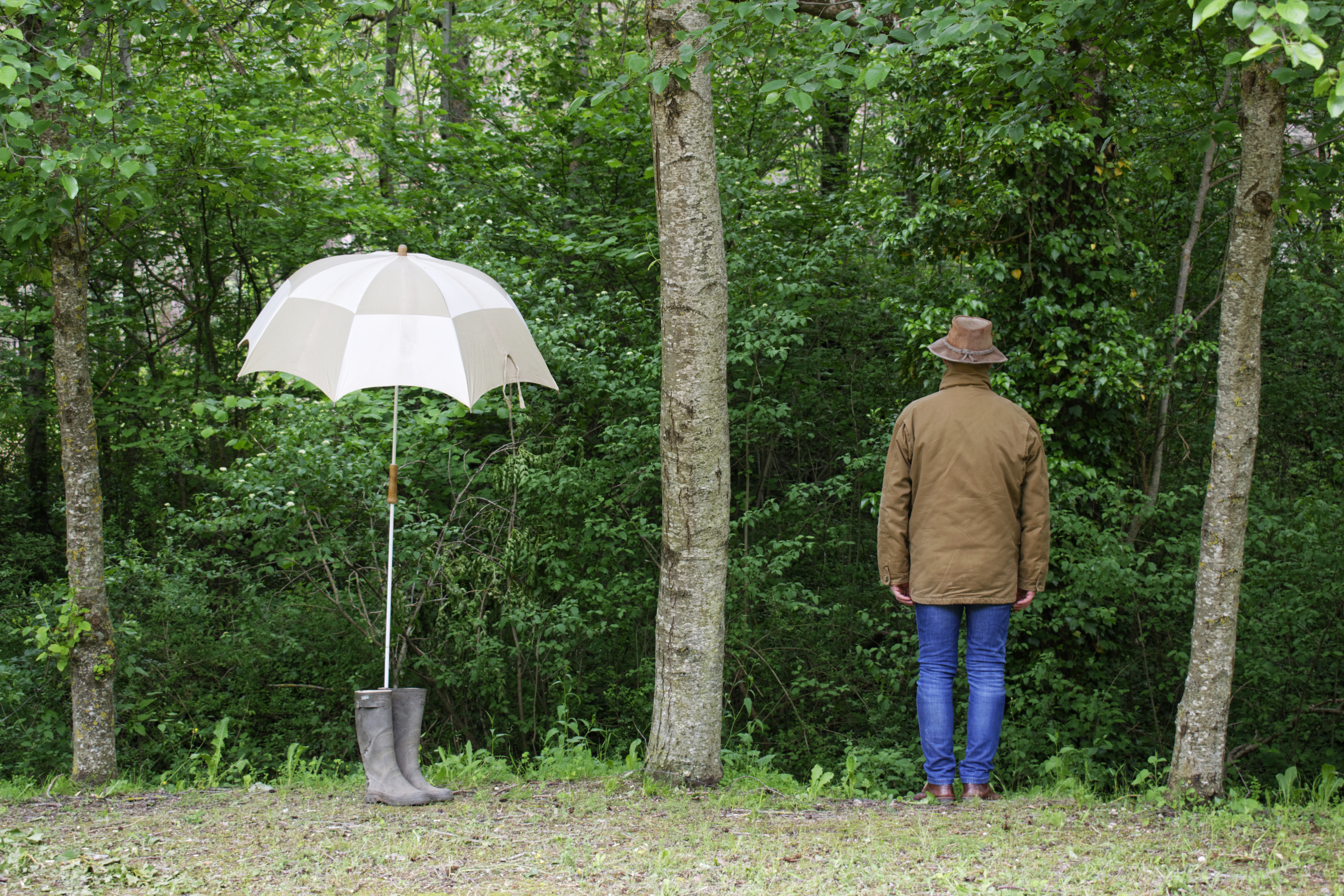 A man standing with his back turned between two trees, to his left a rubber boot carrying a parasol