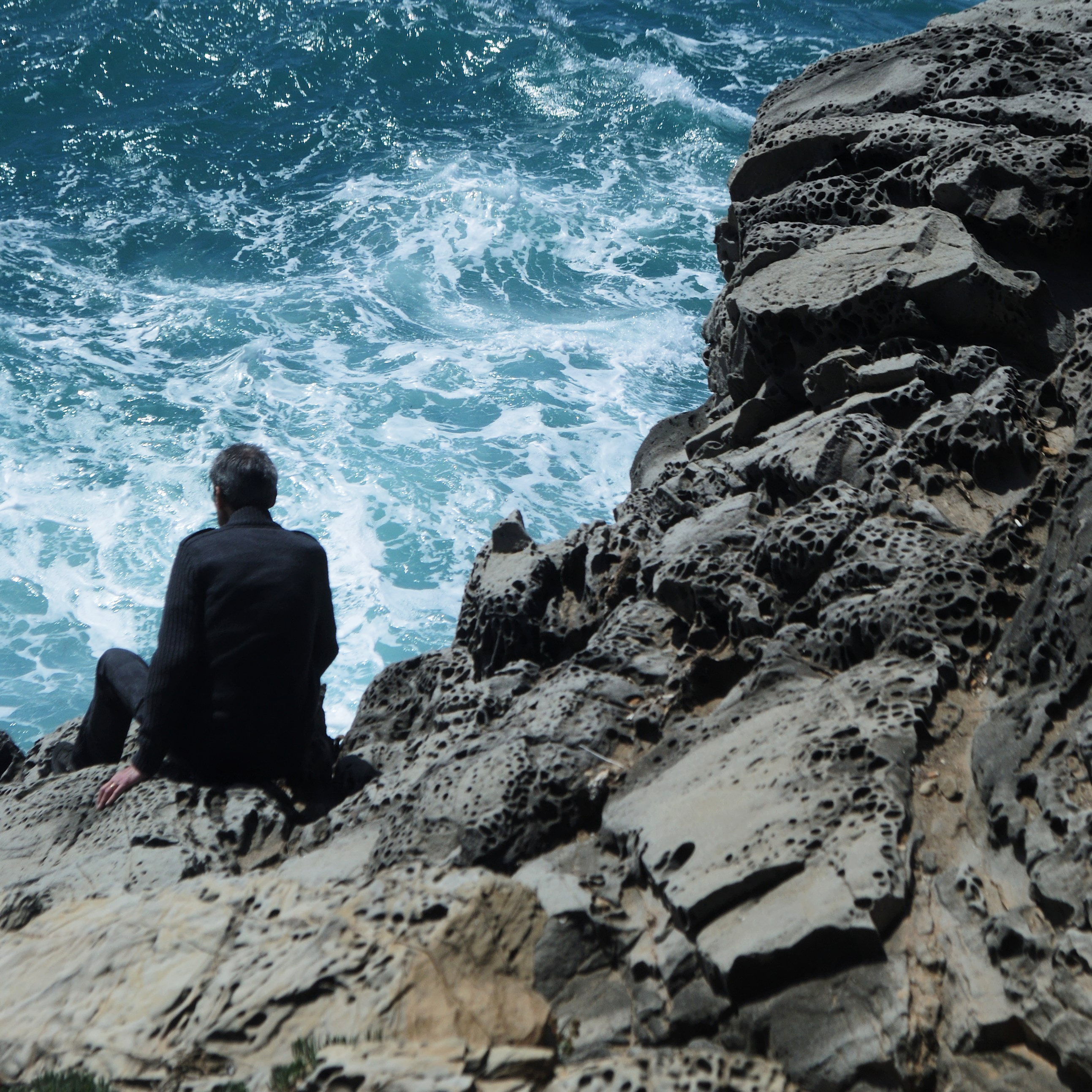 A man in a black suit sitting on the edge of the cliff near the rough sea